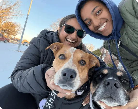Two smiling women and two happy dogs pose together, kneeling side by side.