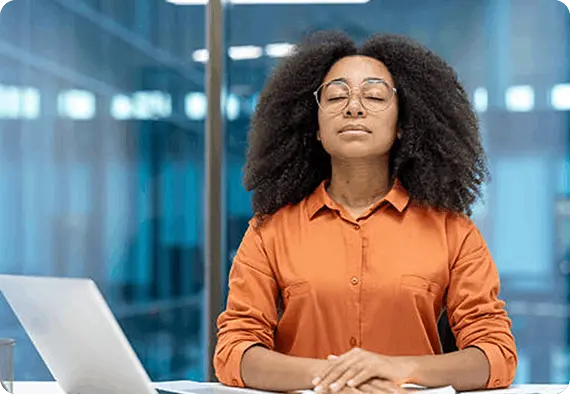 Black woman with glasses meditates at office desk in orange blouse.