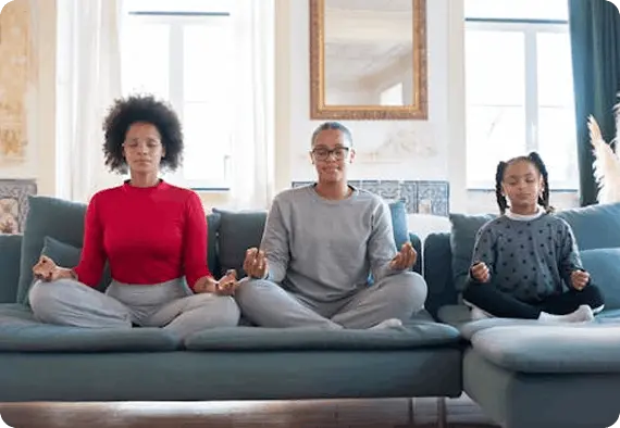 A mother and her two daughters sit cross-legged on a couch, meditating peacefully with eyes shut.